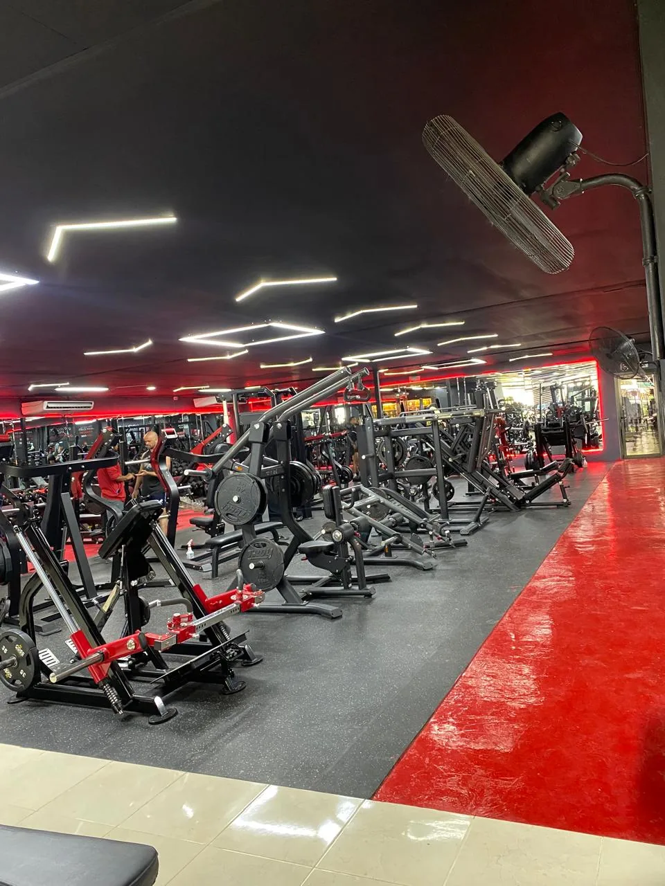 Sala amplia de Best Gym con hileras de máquinas de fuerza, luces geométricas y una franja roja en el piso